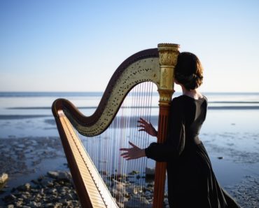 A lady plays her harp while facing the sea.