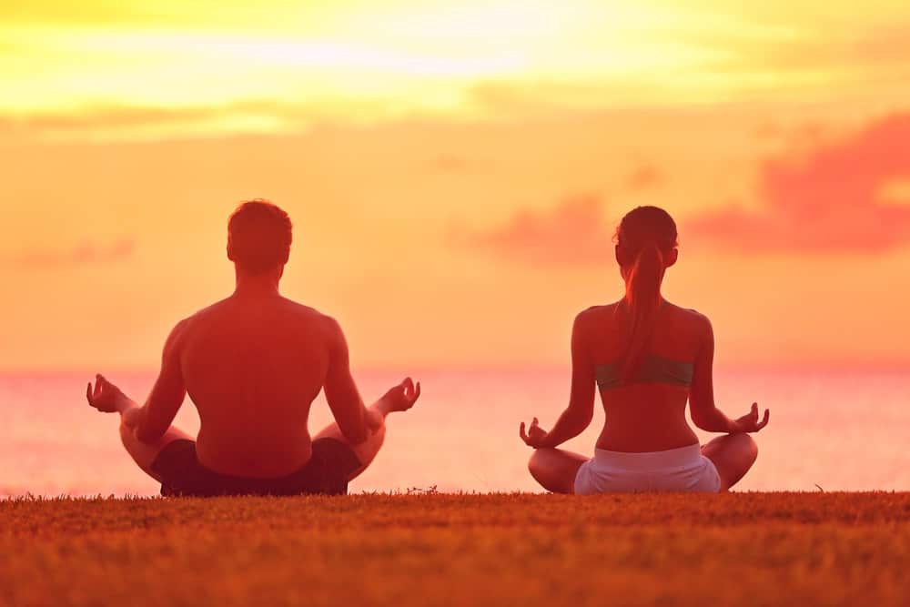 Back view of a man and a woman doing a meditation pose on the ground facing the sunset sky.