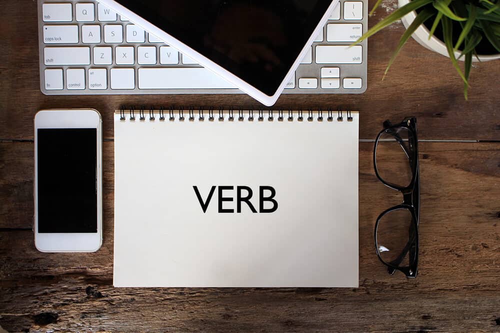The word "verb" written on a notebook placed on a table together with a laptop, a phone and a pair of eyeglasses.
