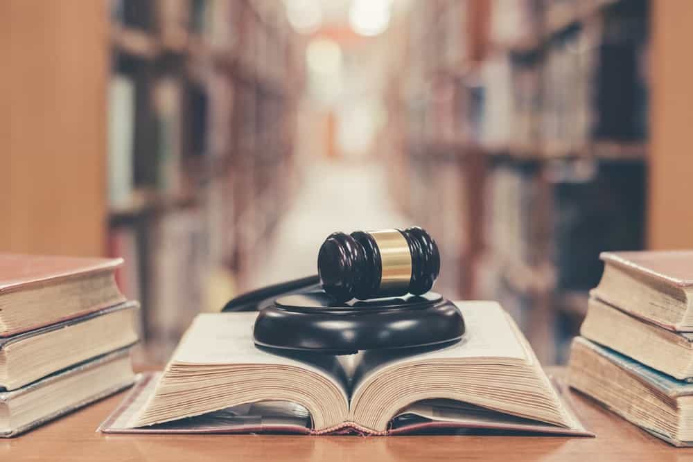 An open book with a judge's black gavel on top is flanked by a trio of stacked hardbound books on wooden desk in a library.