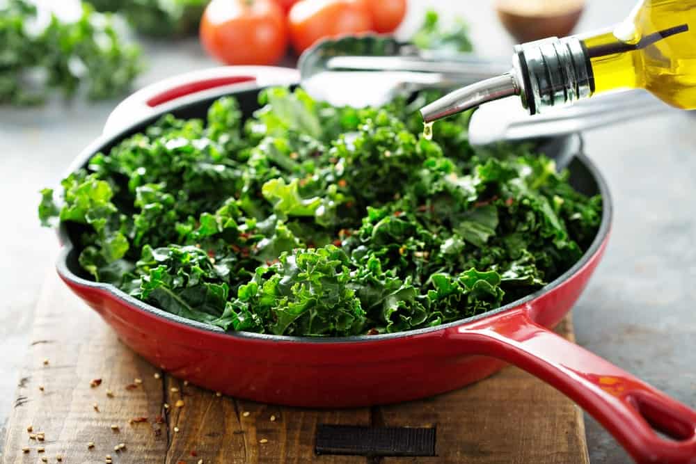 Sprinkling olive oil on kale in a red pan.