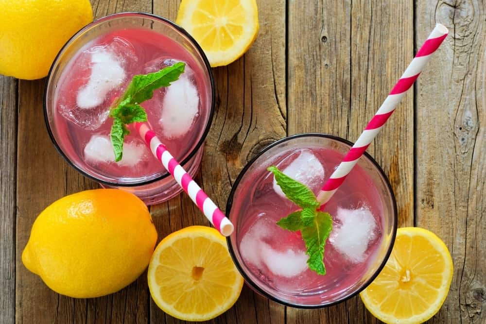 Two glasses of pink lemonade with pink striped straws beside lemons on wooden background.