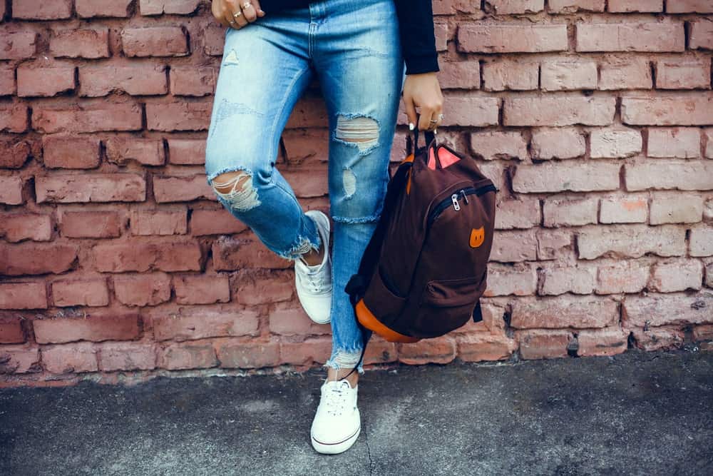 Person in tattered jeans and white sneakers clutching a backpack in one hand while leaning against a brick wall.