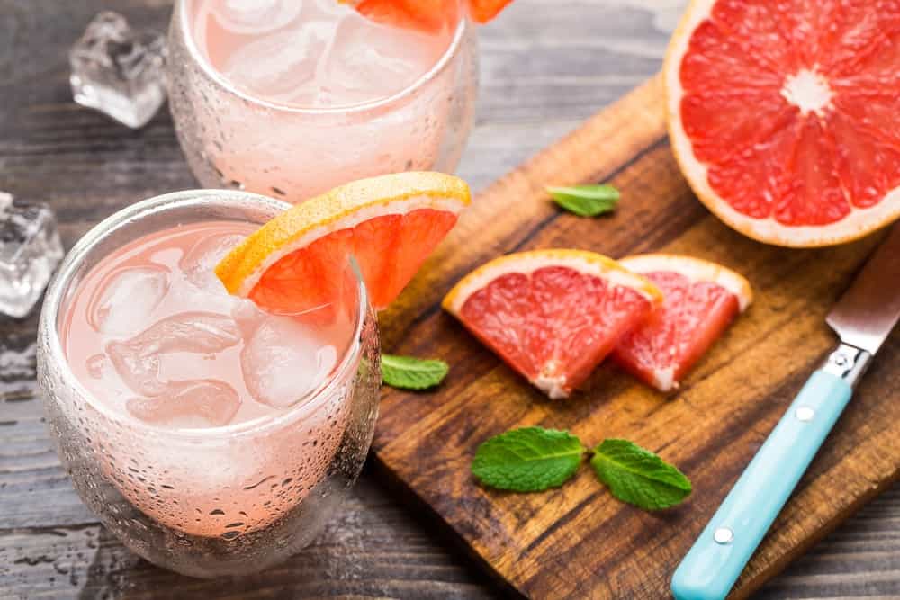 Two glasses of grapefruit cocktail beside a wood chopping board with a knife and slices of grapefruit.