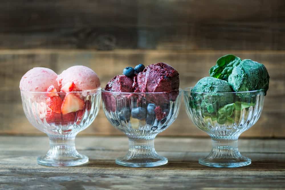 Three glasses of nice cream with flavors of banana, raspberry, and spinach on wooden background.