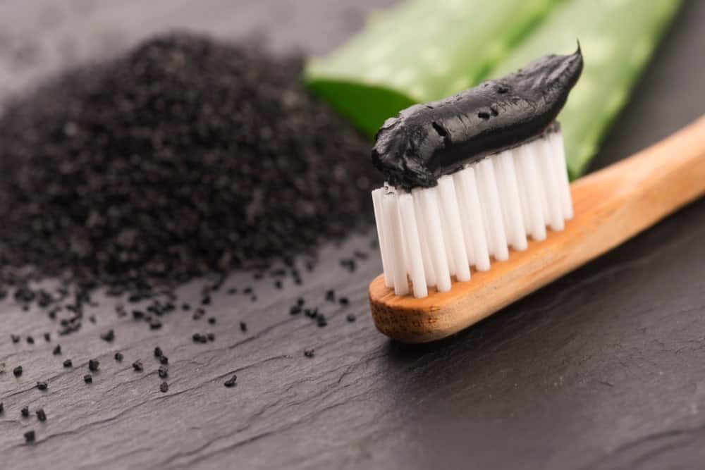 Charcoal and aloe vera toothpaste spread on a bamboo toothbrush beside grounded charcoal and slices of aloe vera.