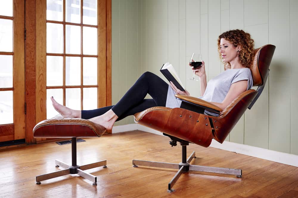 A woman reading a book on an Eames Lounge Chair with a matching footstool.