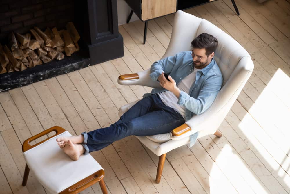 A man browsing his phone while sitting on a lounge chair with foot stool by the fireplace.