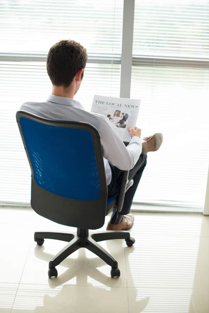 A man reading the newspaper on a swiveling blue office chair with wheels.