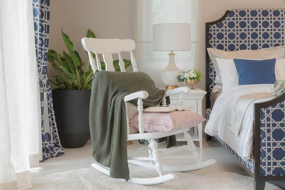 This is a close look at the bedroom with a white wooden rocking chair on the side to act as reading area.