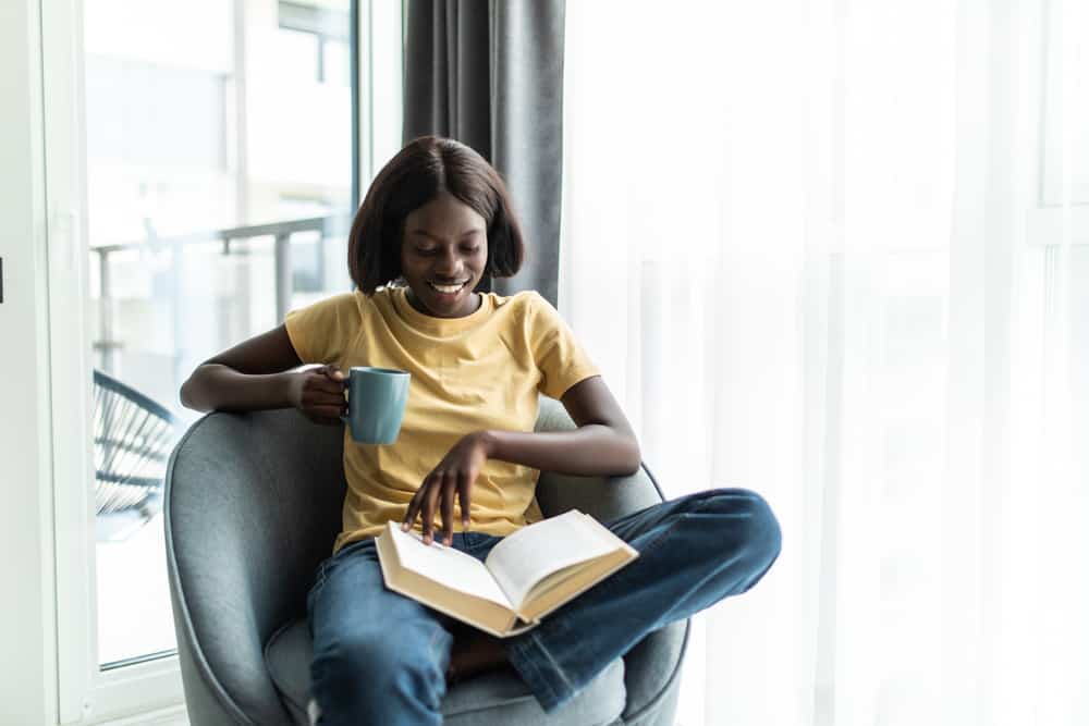 This is a look at a woman reading a book on a barrel chair by the window.